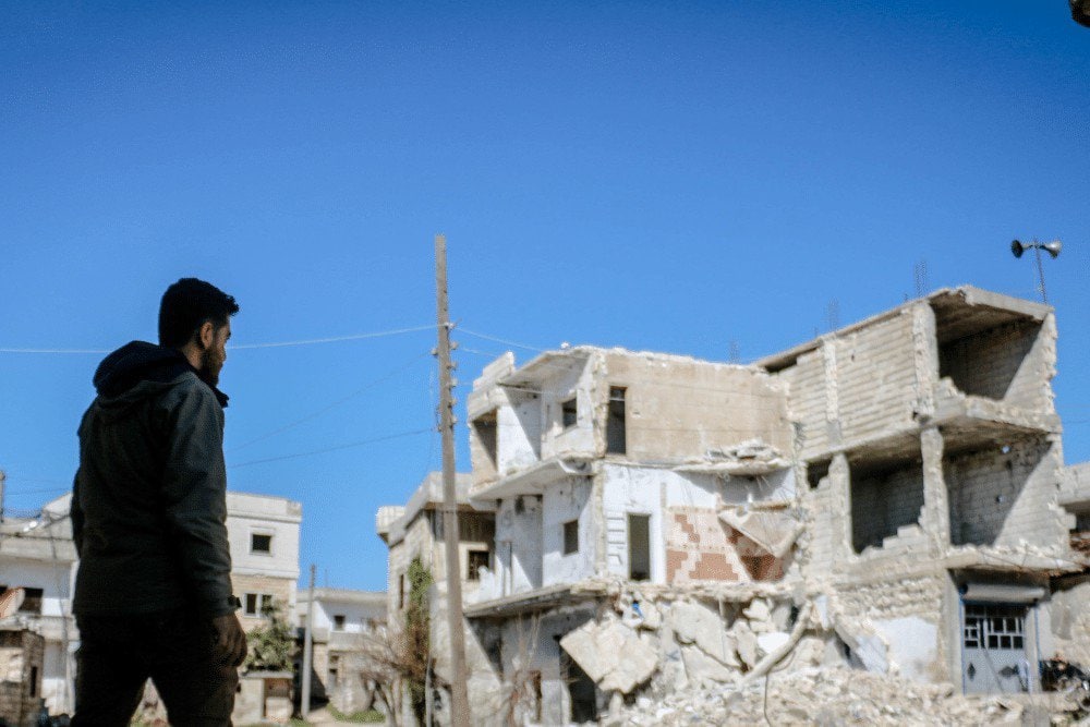 A man stands in front of a destroyed building in syria.